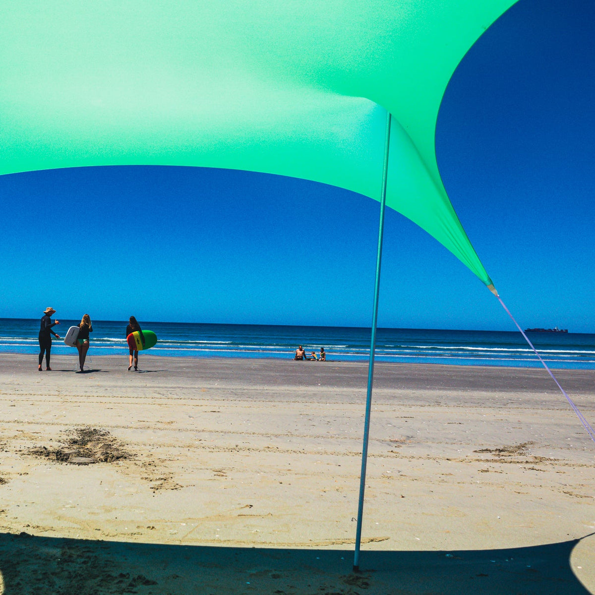 A large mint green tension fabric beach canopy shade structure anchored to a sandy beach, with the ocean and blue sky visible in the background under bright sunlight.